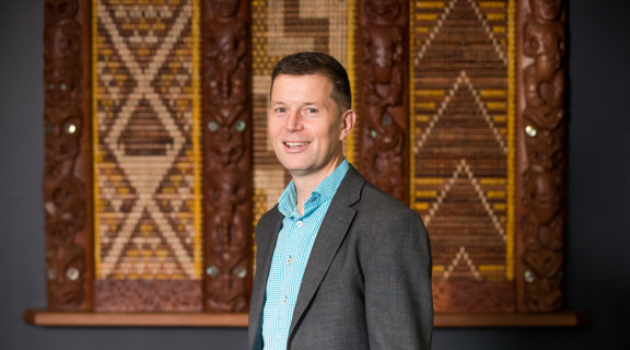 Ben Clark standing in front of a tukutuku panel looking at the camera and smiling