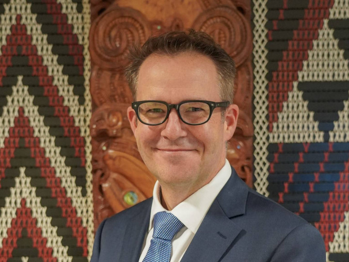 Ben King standing in front of a tukutuku panel looking at the camera and smiling