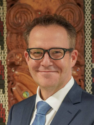 Ben King standing in front of a tukutuku panel looking at the camera and smiling