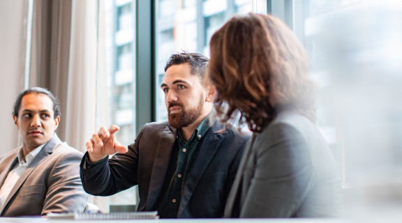 Man wearing suit in a corporate office talking and gesturing with his hand to a group of people