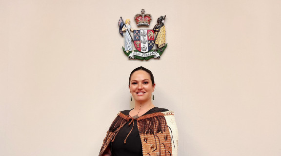 Chloe Graham-Stewart standing below the New Zealand coat of arms looking at the camera and smiling