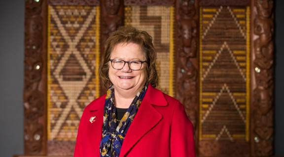 Gloria Campbell standing in front of a tukutuku panel looking at the camera and smiling