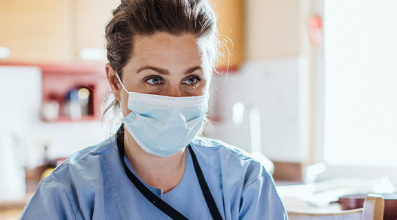 Nurse wearing a medical mask and gloves sitting across the table from an elderly person who is getting a health check-up