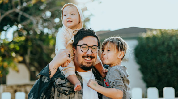 Man and 2 young children standing in front of a white picket fence