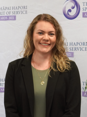 Jacs Bell standing in front of awards banner and smiling at the camera