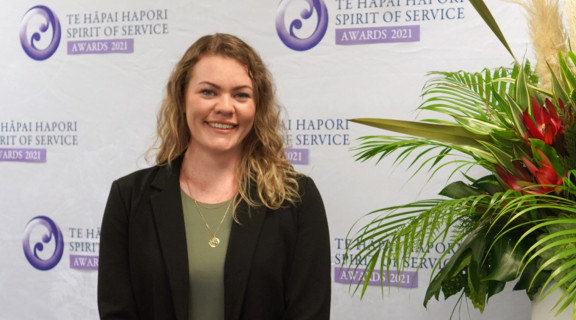 Jacs Bell standing in front of awards banner and smiling at the camera