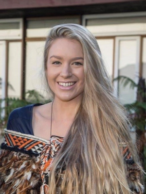 Jesney Te Puke standing outside wearing a kākahu Māori cloak looking at the camera and smiling