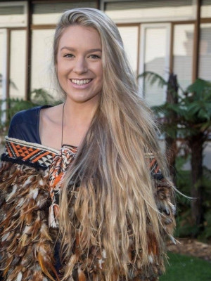 Jesney Te Puke standing outside wearing a kākahu Māori cloak looking at the camera and smiling