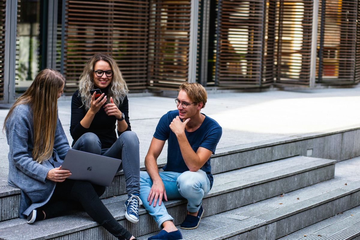 Three people sitting on steps looking at a laptop and talking