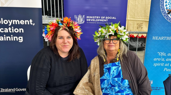 Steph Voight sitting next to a woman, both are wearing flower garlands on their heads and are looking at the camera and smiling