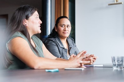 Two women sit at a boardroom table, One is talking to someone not in shot. The other woman is watching her as she speaks.