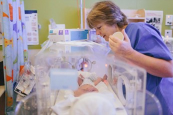 A female nurse in blue scrubs leans over a baby in an incubator. 