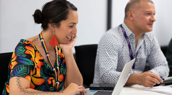 Woman wearing bright colours looking at her laptop and smiling as she sits next to a colleague in a meeting room.