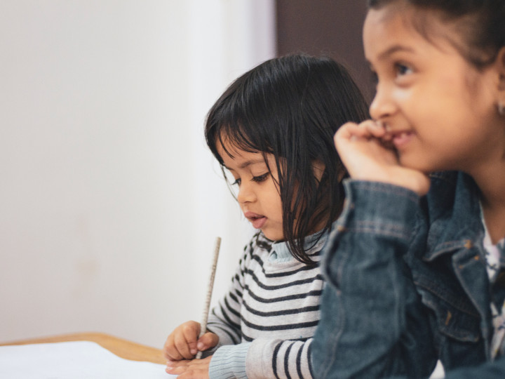 Two young children sitting next to each other at a desk, one is using a pencil to write