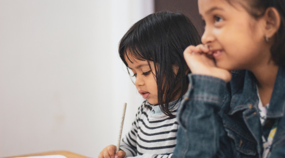 Two young children sitting next to each other at a desk, one is using a pencil to write