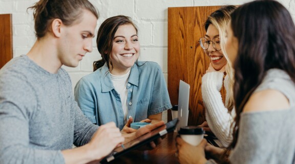 Four people sitting around a wooden table, engaged in discussion and smiling. One person is writing on a tablet, another holds a cup, and a laptop is open on the table. The setting appears casual and collaborative.