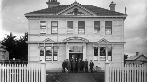 Government department building in Stratford with employees standing outside in 1913. The windows advertise the following: Registrar of Brands, Inspector of Stock, Department of Agriculture - Industries & Commerce, Inspector of Noxious weeds, Land & Survey