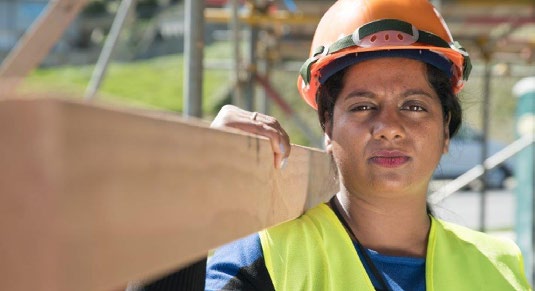 A woman is working on a building site. She is wearing a hard hat and high vis vest, and carrying a plank of wood over one  shoulder.
