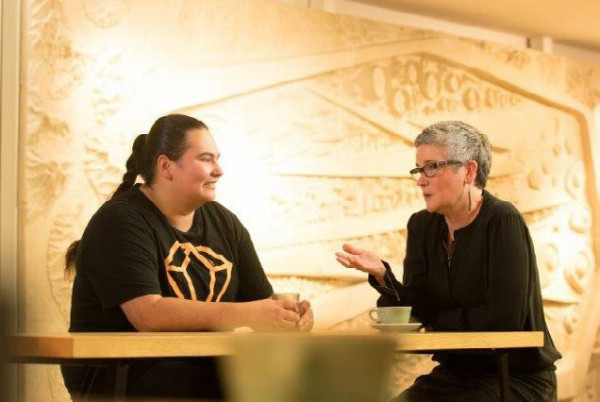 Two women sit at a table in a cafe, with cups in front of them on the table. The woman on the right is talking, while the woman on the left listens and smiles.