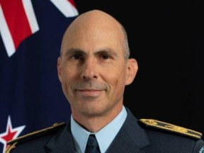 Andrew Clark wearing uniform standing in front of the New Zealand flag and smiling at the camera