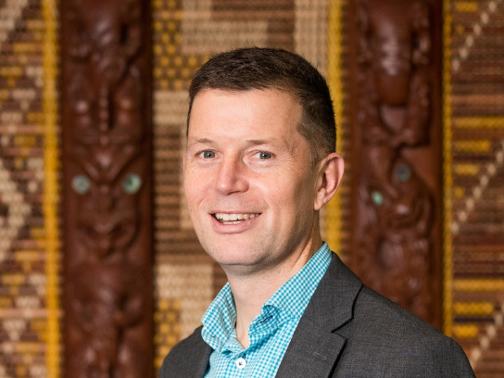 Ben Clark standing in front of a tukutuku panel looking at the camera and smiling