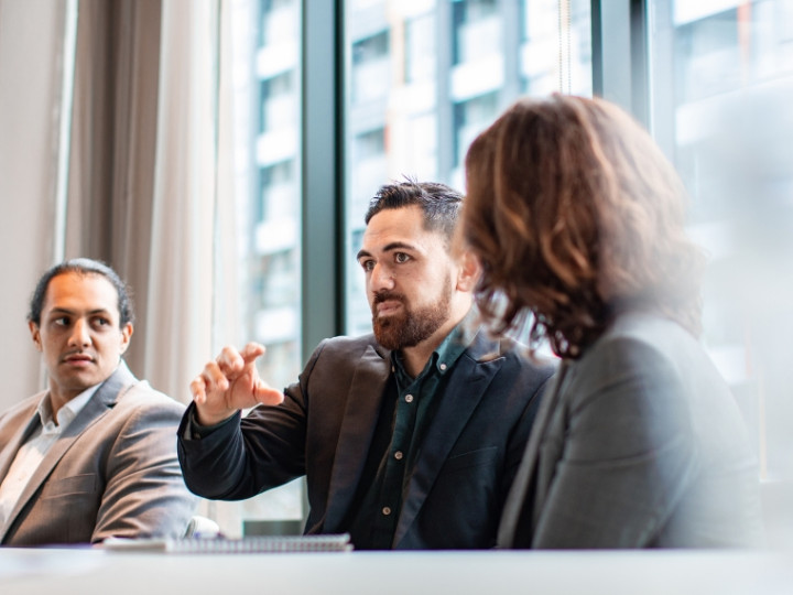 Man wearing suit in a corporate office talking and gesturing with his hand to a group of people