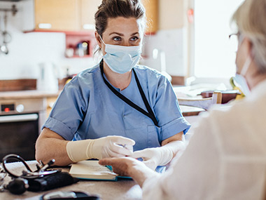 Nurse wearing a medical mask and gloves sitting across the table from an elderly person who is getting a health check-up