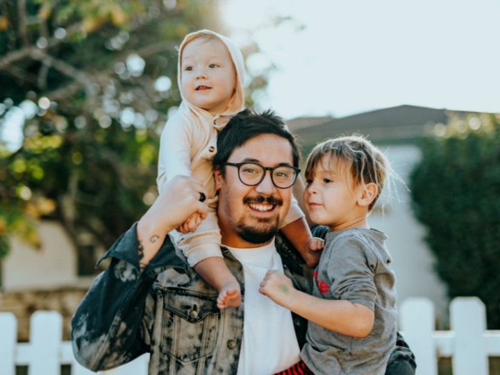 Man and 2 young children standing in front of a white picket fence