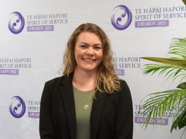 Jacs Bell standing in front of awards banner and smiling at the camera