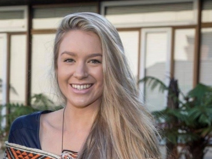Jesney Te Puke standing outside wearing a kākahu Māori cloak looking at the camera and smiling