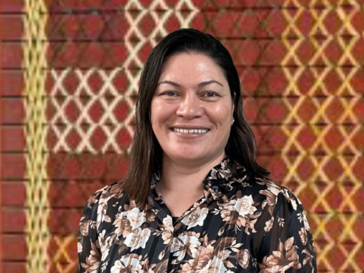 Jules Lynch standing in front of a tukutuku panel looking at the camera and smiling