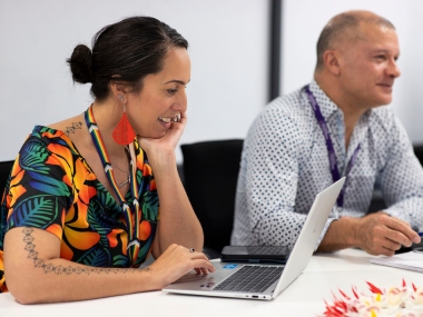 Woman wearing bright colours looking at her laptop and smiling as she sits next to a colleague in a meeting room.